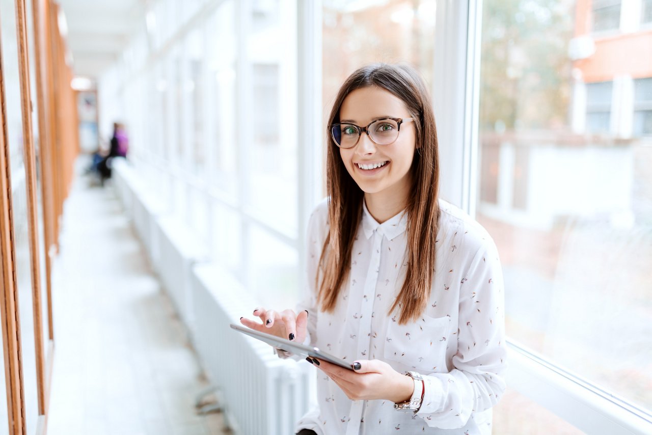 Smiling Caucasian collage girl using tablet while standing in front of noticeboard and looking at camera. University hall interior.