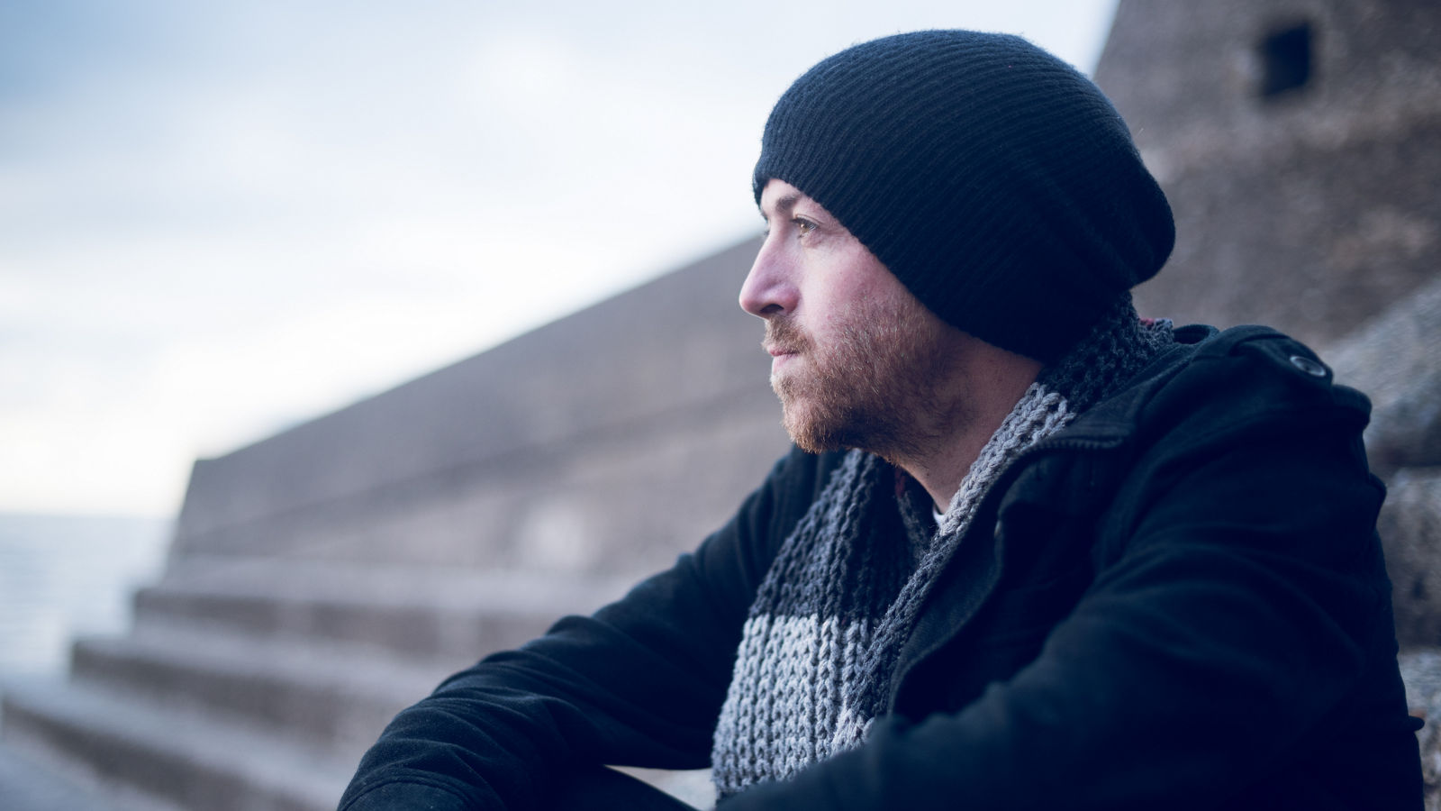 A young handsome man sitting alone by steps and contemplating life by the quiet sea, surrounded by raw concrete sea defence wall. Mental health and depression road to recovery.