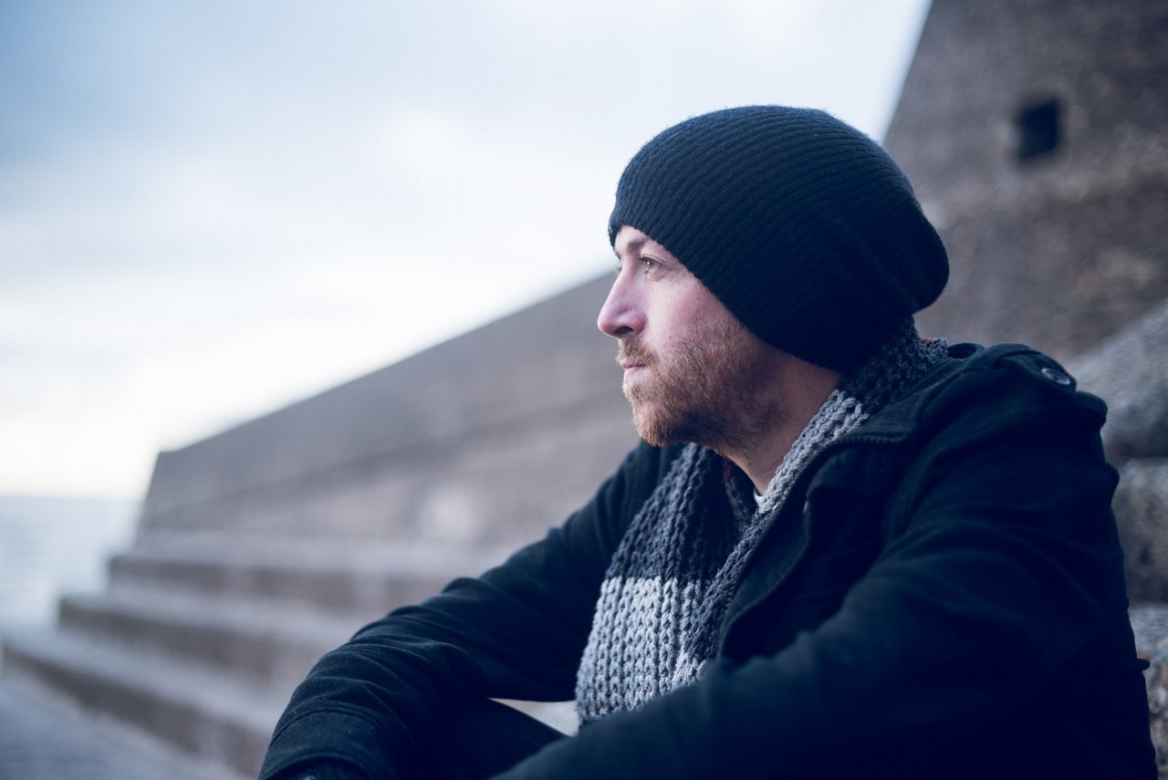 A young handsome man sitting alone by steps and contemplating life by the quiet sea, surrounded by raw concrete sea defence wall. Mental health and depression road to recovery.