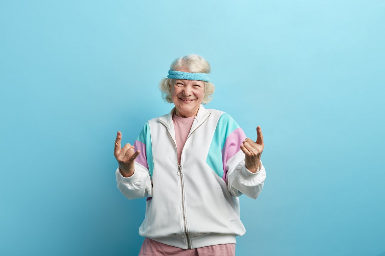 Cute hipster grandmother smiling and making rock sign against blue studio background
