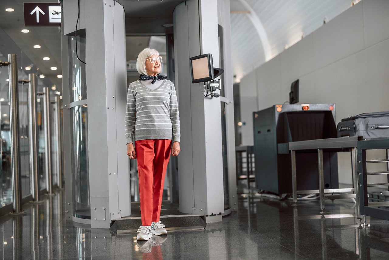 Full length portrait of old lady in glasses waiting for passing through the metal detector frame before flight