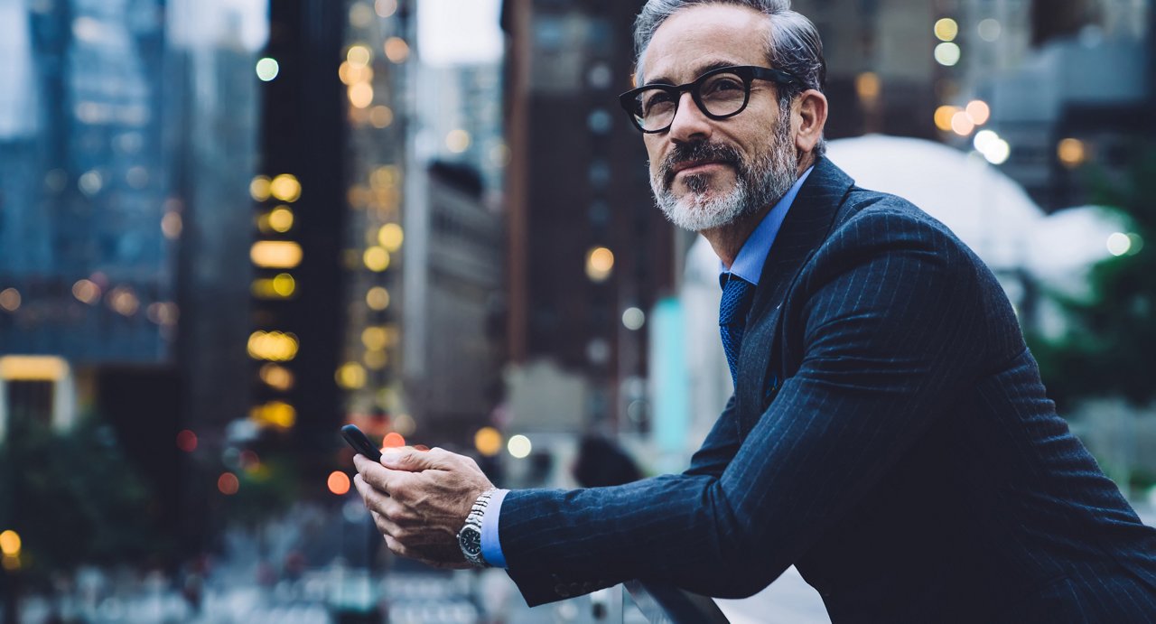 Middle aged man in glasses and business suit holding phone and leaning on glass fence against background of New York street at evening
