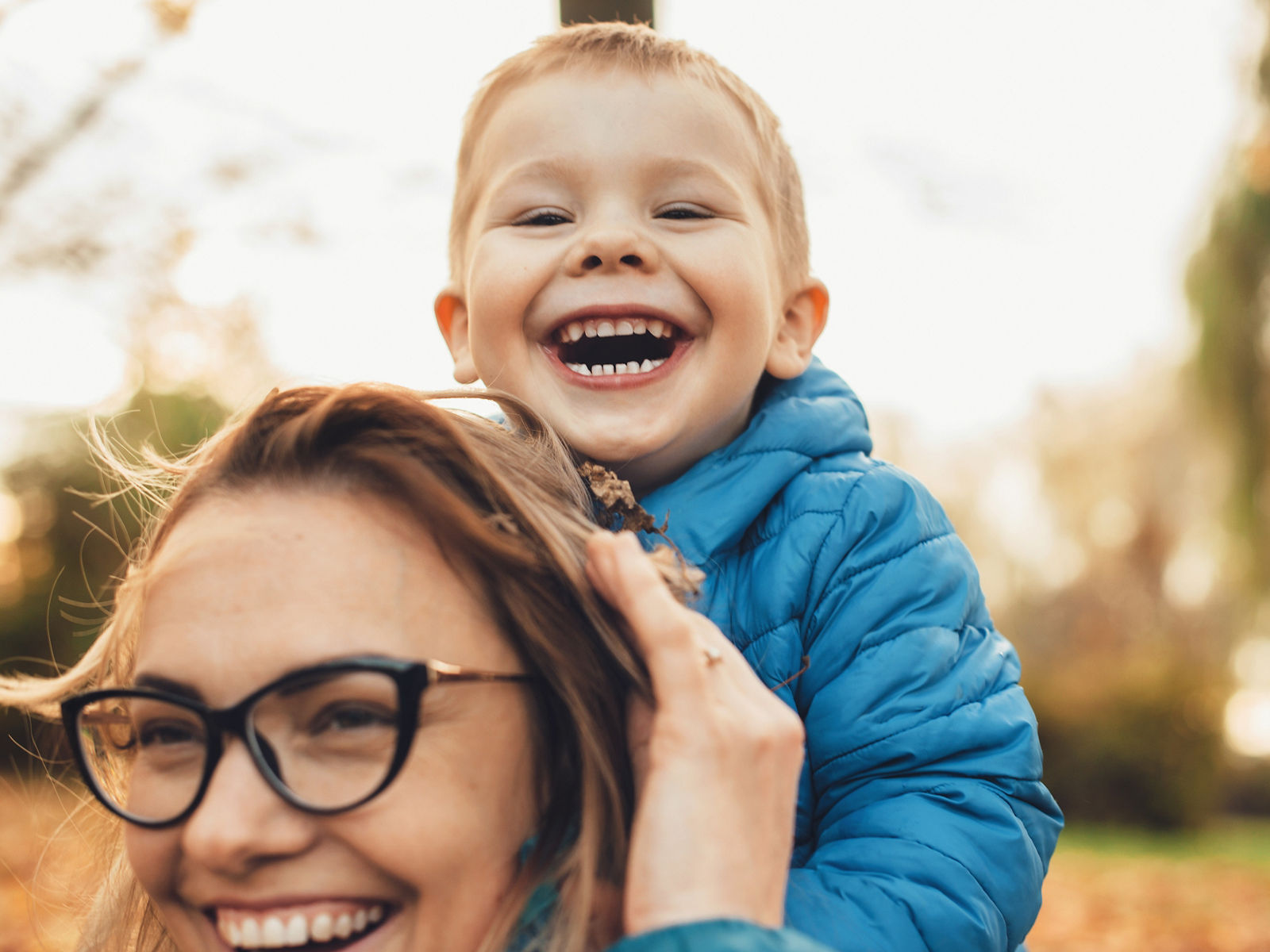 Lovely little kid laughing while sitting on his mother's back outdoor looking at camera.