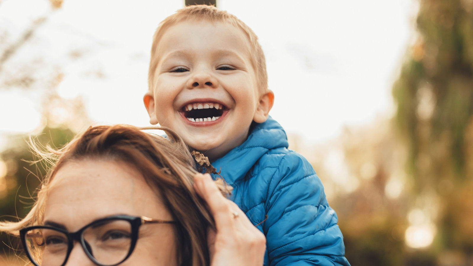 Lovely little kid laughing while sitting on his mother's back outdoor looking at camera.