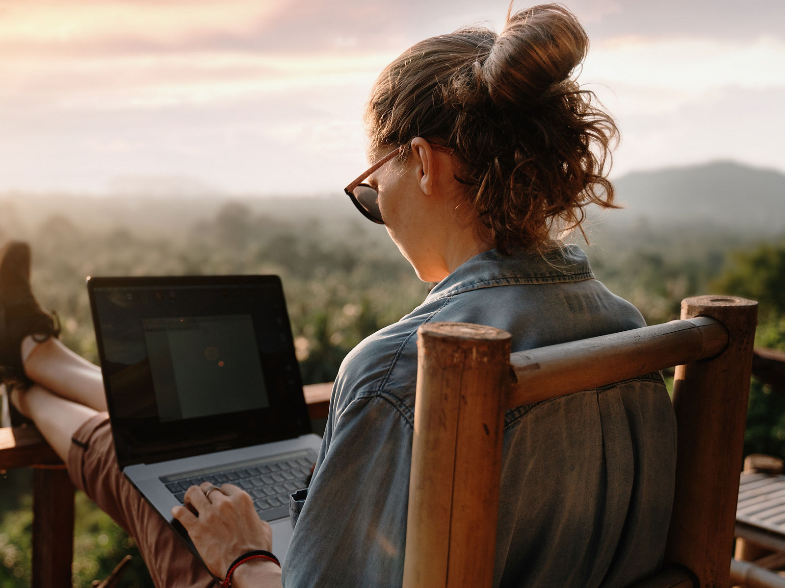 Eine junge Frau sitzt auf einem Stuhl mit hochgelegten Füßen, auf einer Terrasse sitzend arbeitet sie am Laptop, mit Ausblick auf weite Natur