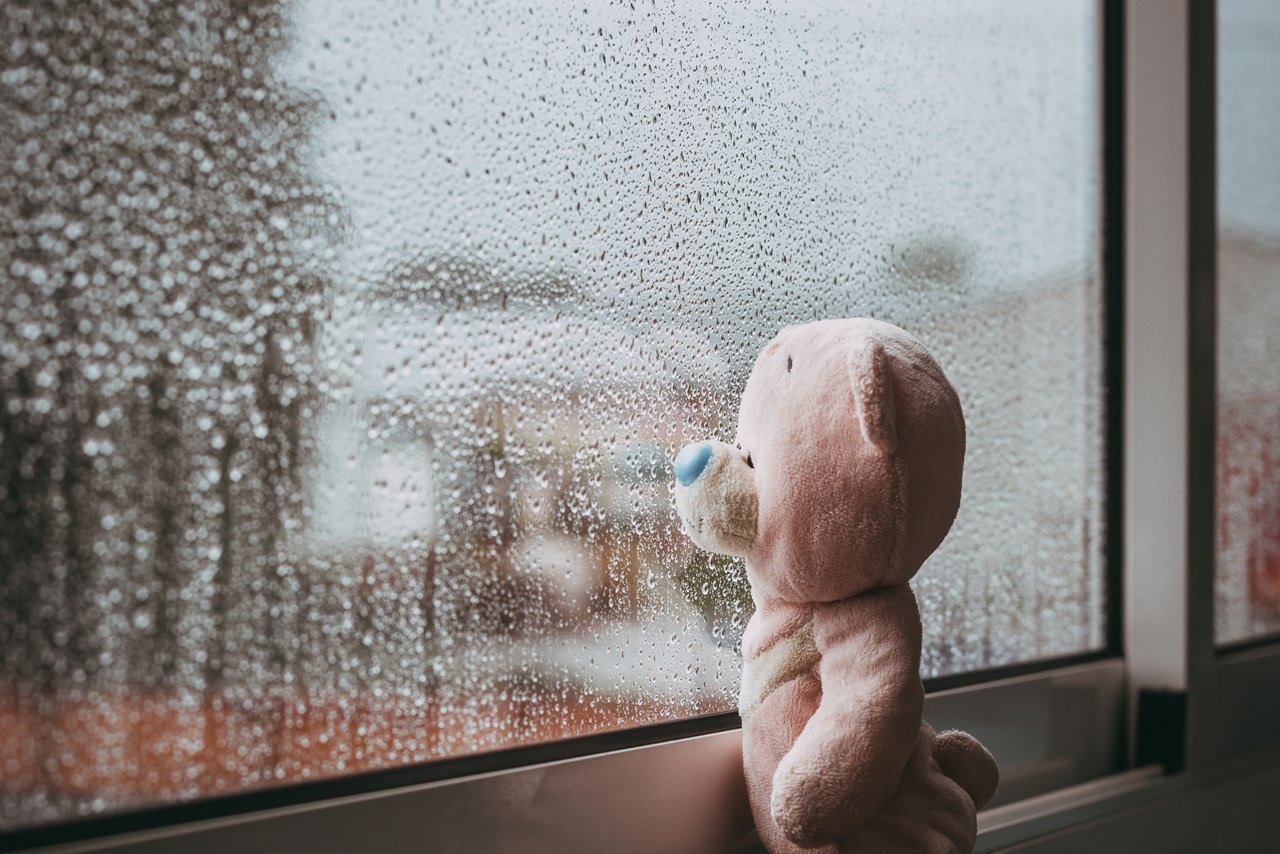 Ein trauriger Spielzeugbär schaut aus dem Fenster an einem regnerischen Herbsttag. Regentropfen auf dem Fenster.