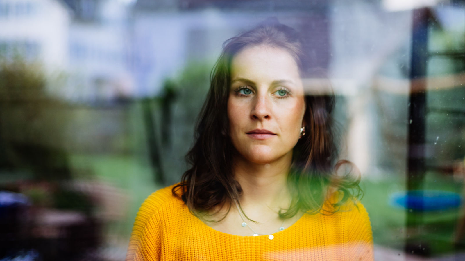 Young woman looks thoughtfully and sadly through the window into the garden with children's toys