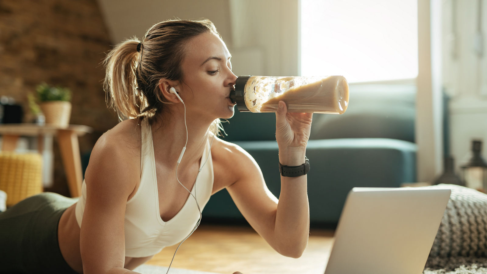 Young athletic woman drinking protein shake while using laptop on the floor. 