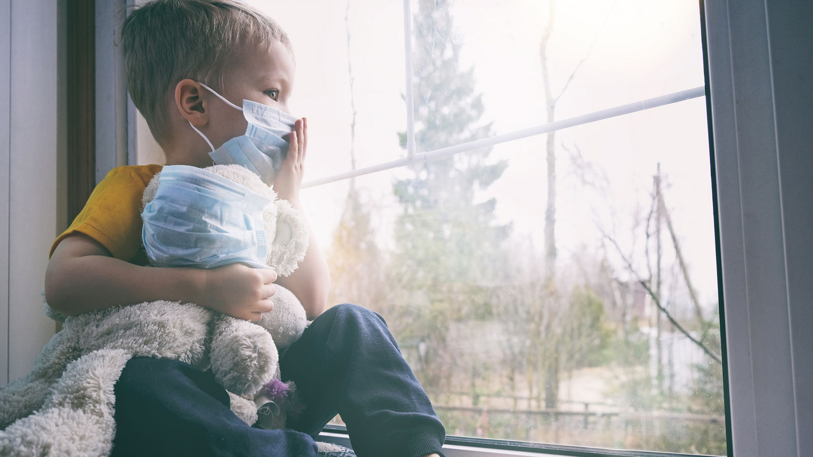 Illness child on home quarantine. Boy and his teddy bear both in protective medical masks sits on windowsill and looks out window. Virus protection, coronavirus pandemic, prevention epidemic.