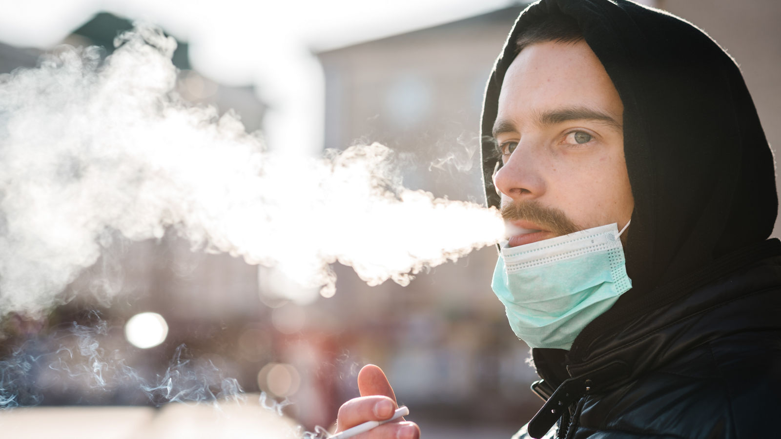 Smoking. Closeup man with mask during COVID-19 pandemic smoking a cigarette at the street. Smoking causes lung cancer and other diseases. The dangers and harm of smoking. Coronavirus.,Smoking. Closeup man with mask during COVID-19 pandemic smoking 