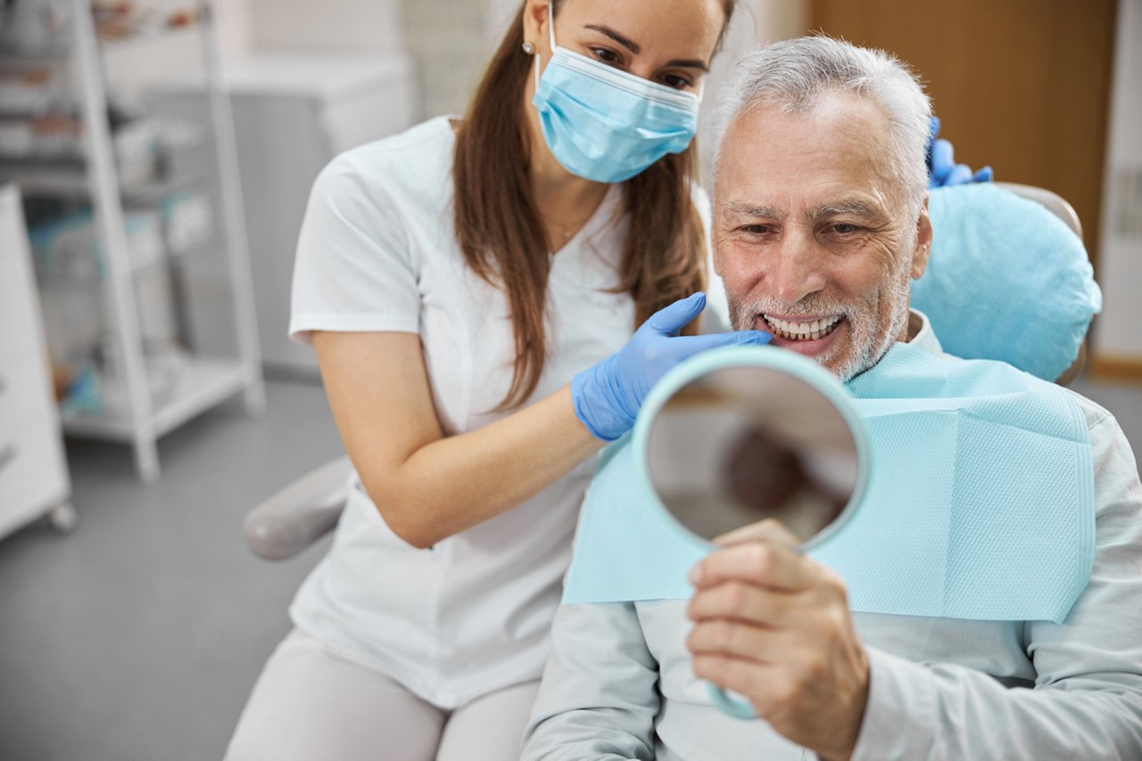 Elderly man and his young female dentist smiling while looking in the mirror in dental clinic