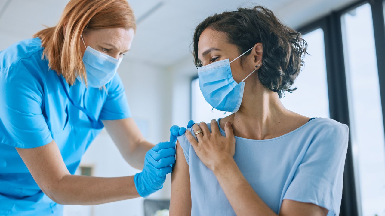 Medical Nurse in Safety Gloves and Protective Mask is Making a Vaccine Injection to a Female Patient in a Health Clinic. Doctor Uses Hypodermic Needle and a Syringe to Put a Shot of Drug as Treatment.