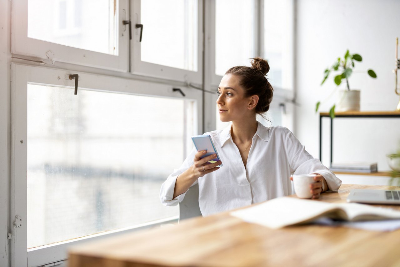 Eine junge Frau hält ein Smartphone und eine Tasse in der Hand, schaut aus dem Fenster und sitzt vor dem Laptop am Schreibtisch.