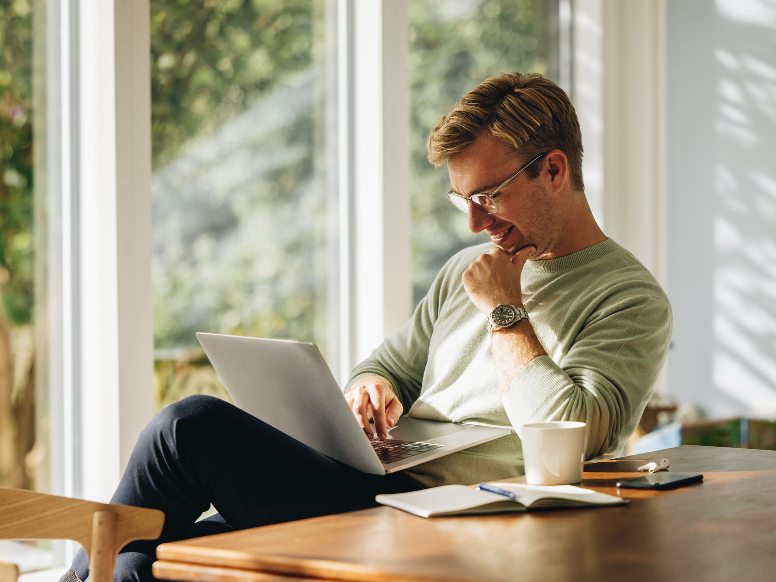 Young man using laptop and smiling at home. Man sitting by table working on laptop computer.