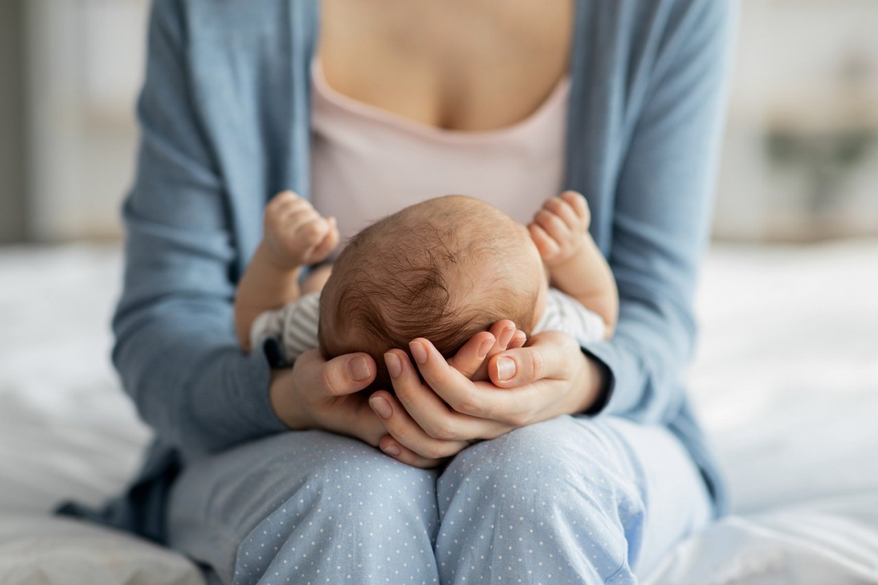 Mother Care. Unrecognizable mom holding newborn baby on her laps, lulling him to sleep, loving mum bonding with her infant child at home, cropped image with selective focus