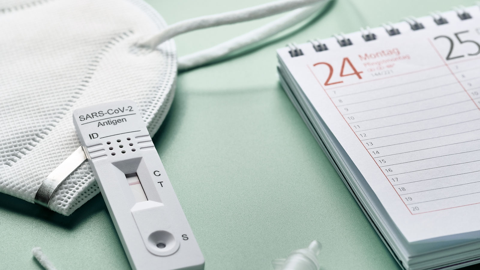 Week planner, chnelltest, rapid corona test in German language. Week planner calendar and pp2 medical face mask on green table. Test-to-go concept.
