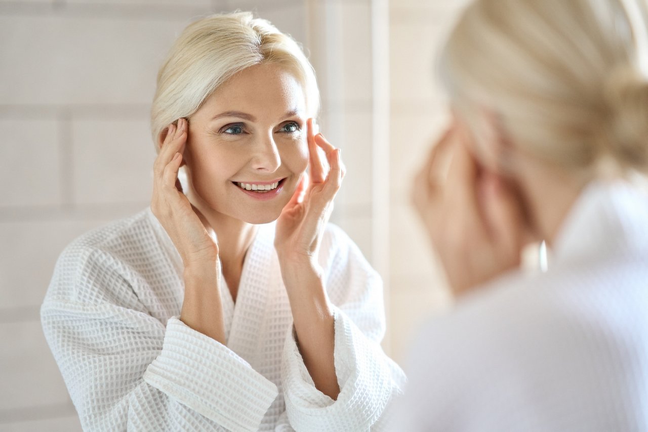 Gorgeous mid age adult 50 years old blonde woman standing in bathroom after shower touching face, looking at reflection in mirror smiling doing morning beauty routine. Older dry skin care concept.