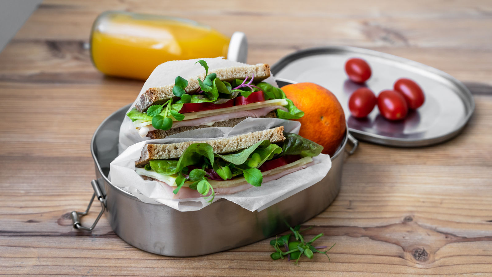 Sandwich mit Salatblätter und Schinken in einer wiederverwendbaren Blechdose, Orangensaft und Cocktailtomaten auf einem Holz Tisch.