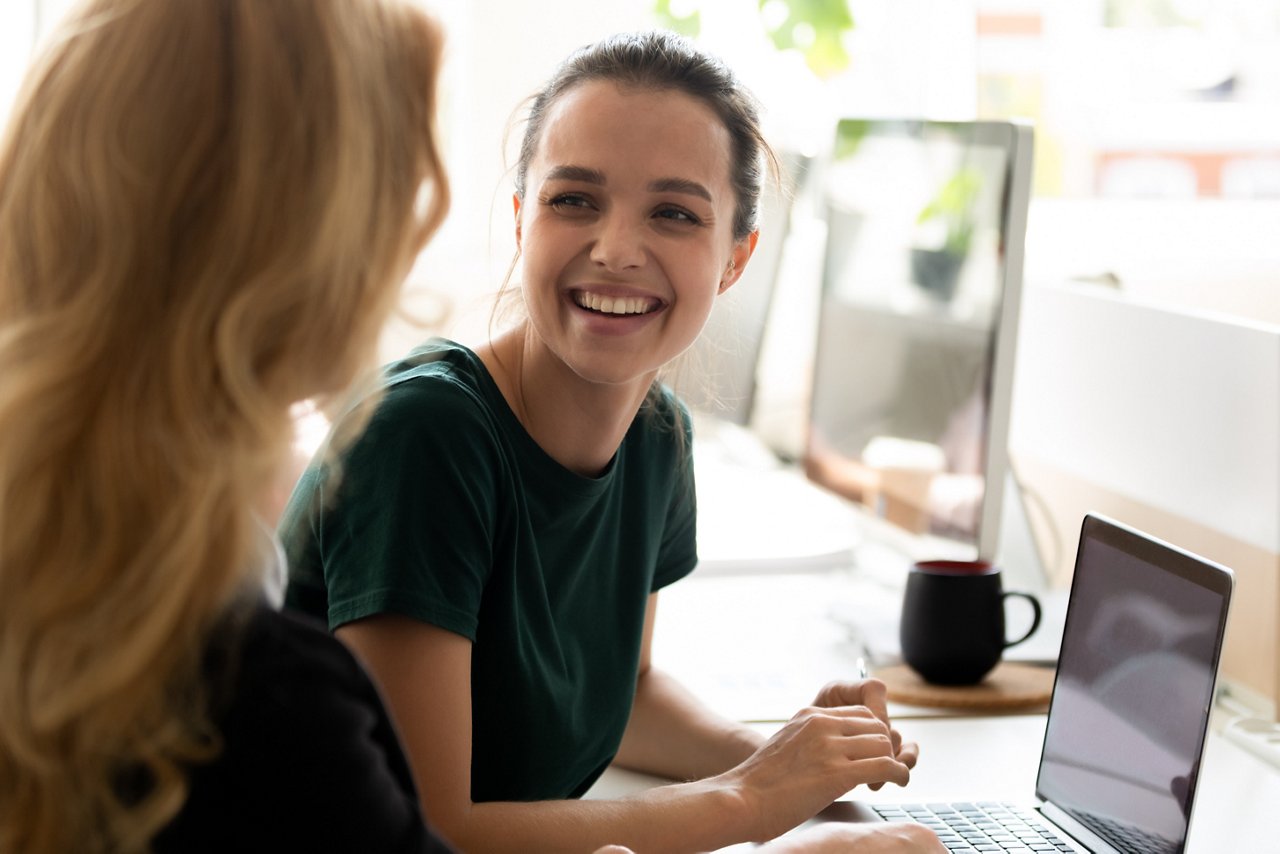 Happy millennial intern, student girl talking to teacher, consulting tutor, mentor at computer in training center, discussing new professional skills, smiling, laughing. Business education, internship