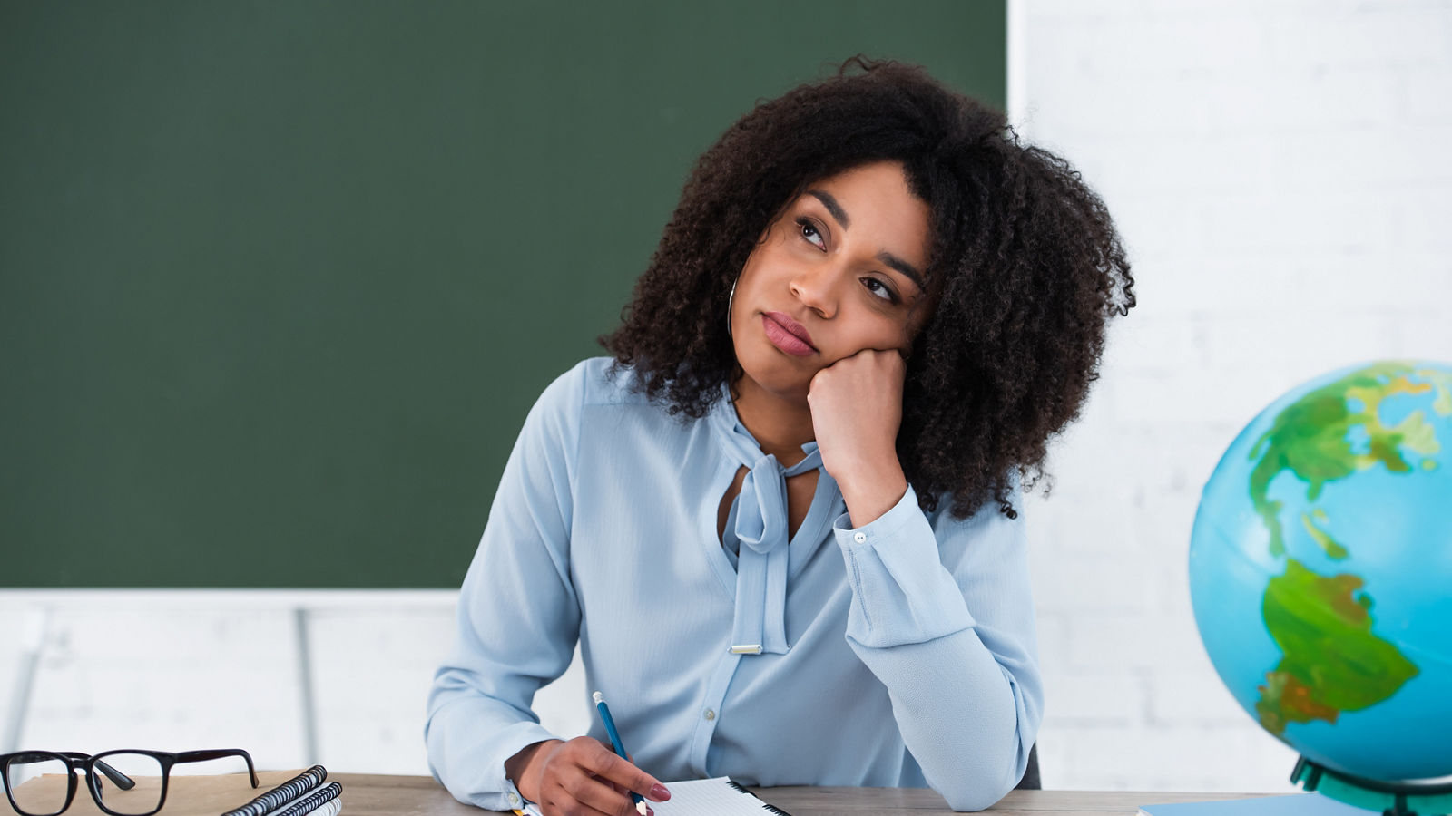 Pensive african american teacher sitting near stationery, eyeglasses and globe in classroom