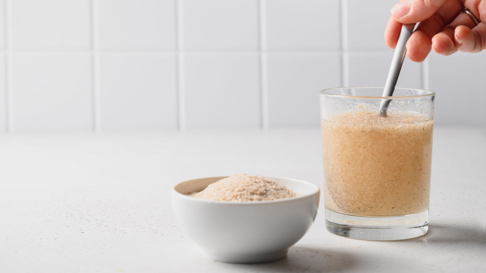 Woman adds spoon psyllium fiber and mix up in glass of water on a kitchen white background. Superfood for healthy intestines and gluten free diet.