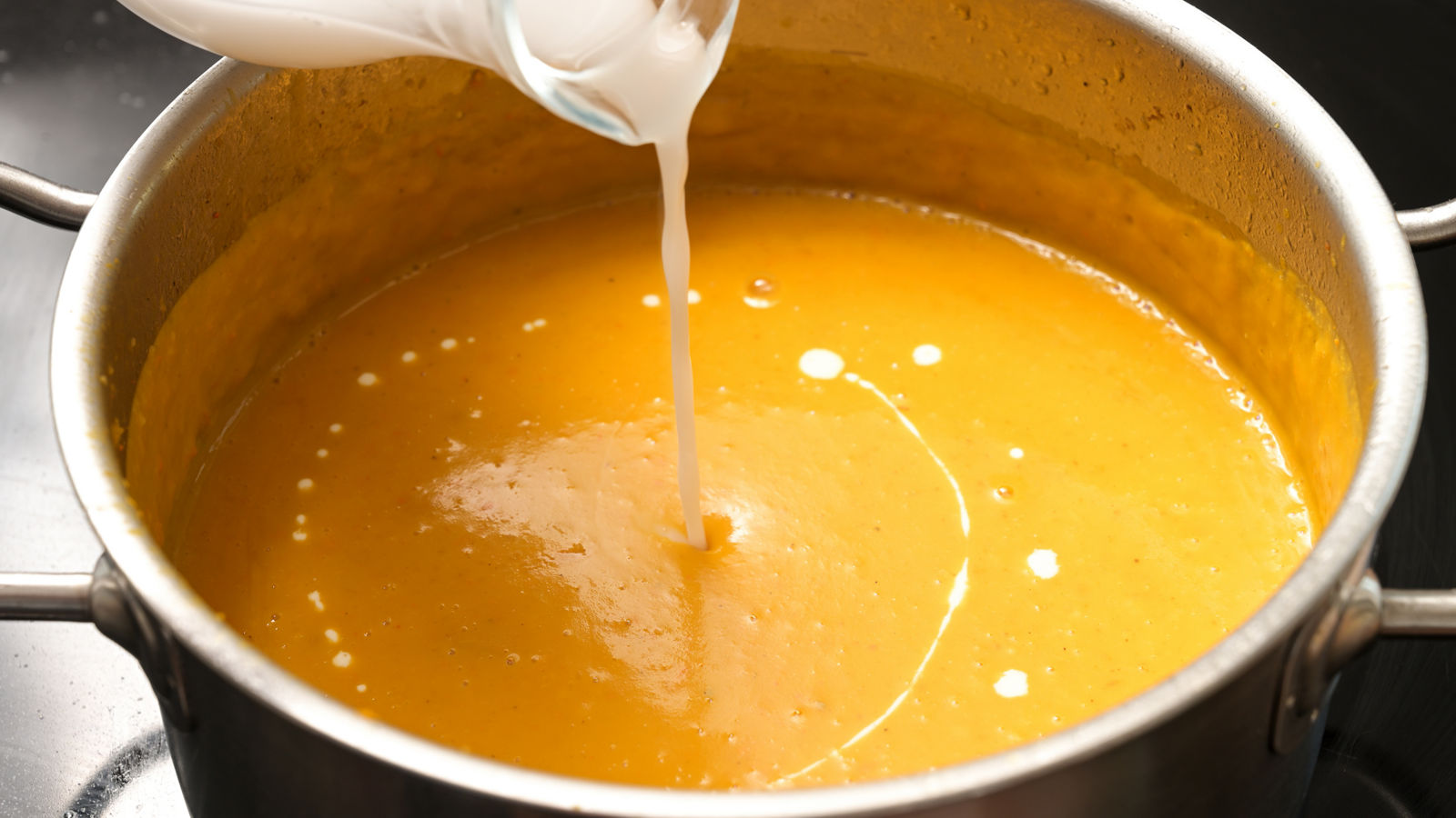 Pouring coconut milk into a vegetable cream soup from squash in a steel pot, vegan cooking in autumn, copy space, selected focus