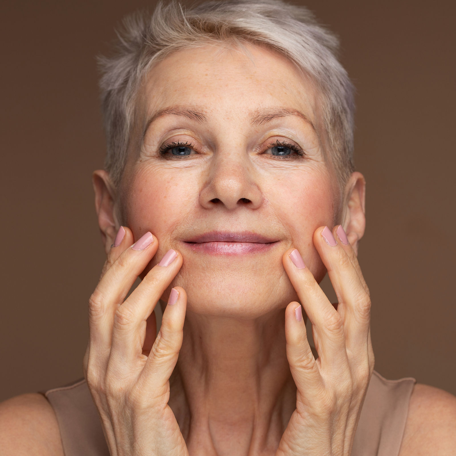 Portrait of gorgeous happy mature woman, senior older 60 year lady looking at camera touching her face isolated on brown background. Ads of lifting anti wrinkle skin care, spa. Copy space.