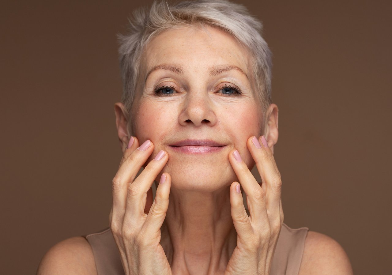 Portrait of gorgeous happy mature woman, senior older 60 year lady looking at camera touching her face isolated on brown background. Ads of lifting anti wrinkle skin care, spa. Copy space.