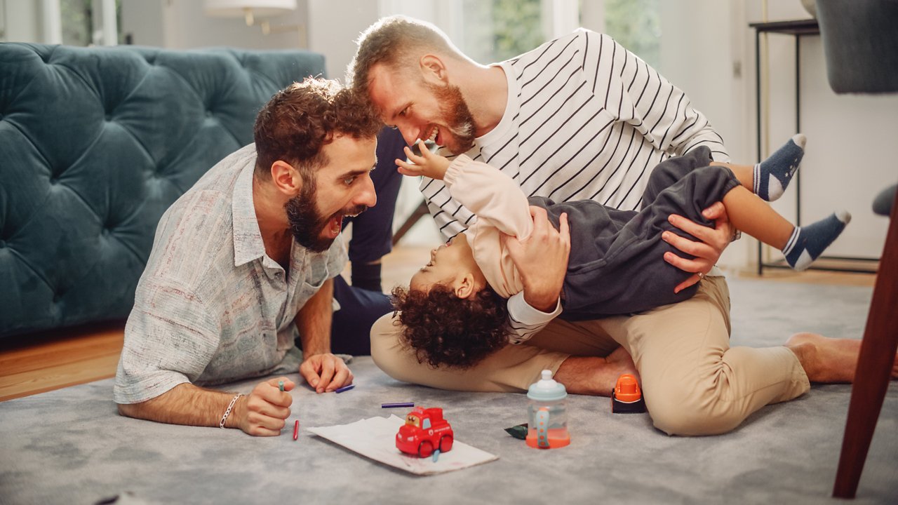 Loving LGBTQ Family Playing with Toys with Adorable Baby Boy at Home on Living Room Floor. Cheerful Gay Couple Nurturing a Child. Concept of Diverse Childhood, New Life, Parenthood.