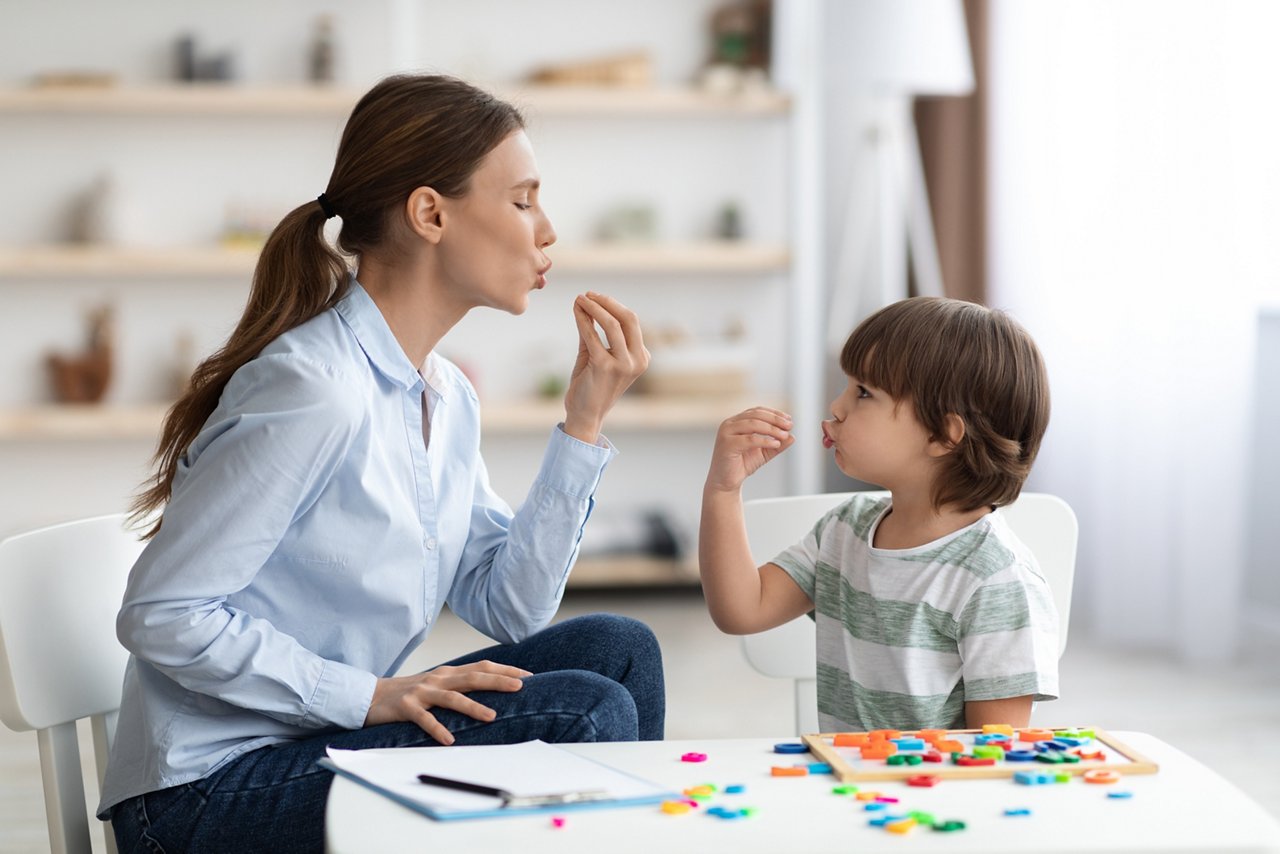 Young woman speech therapist studying together with small kid, learning practice pronunciation exercises with little boy child at office, free space