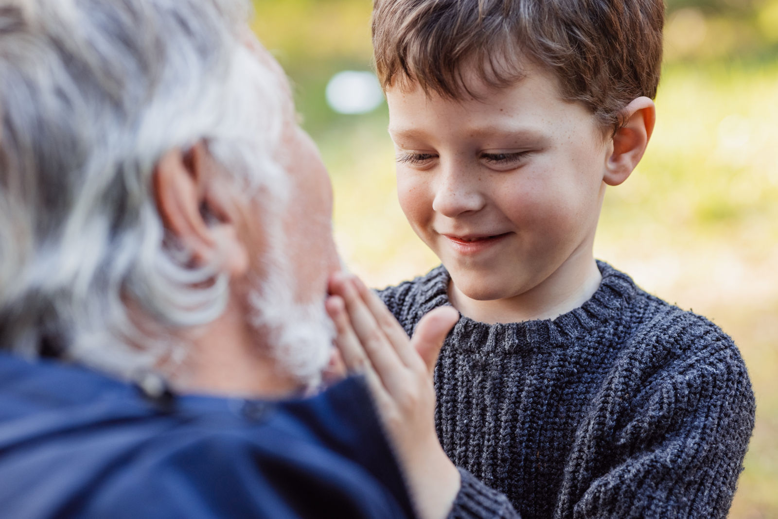 Cheerful little grandson in warm sweater touching gray beard of smiling senior grandfather with closed eyes on blurred background