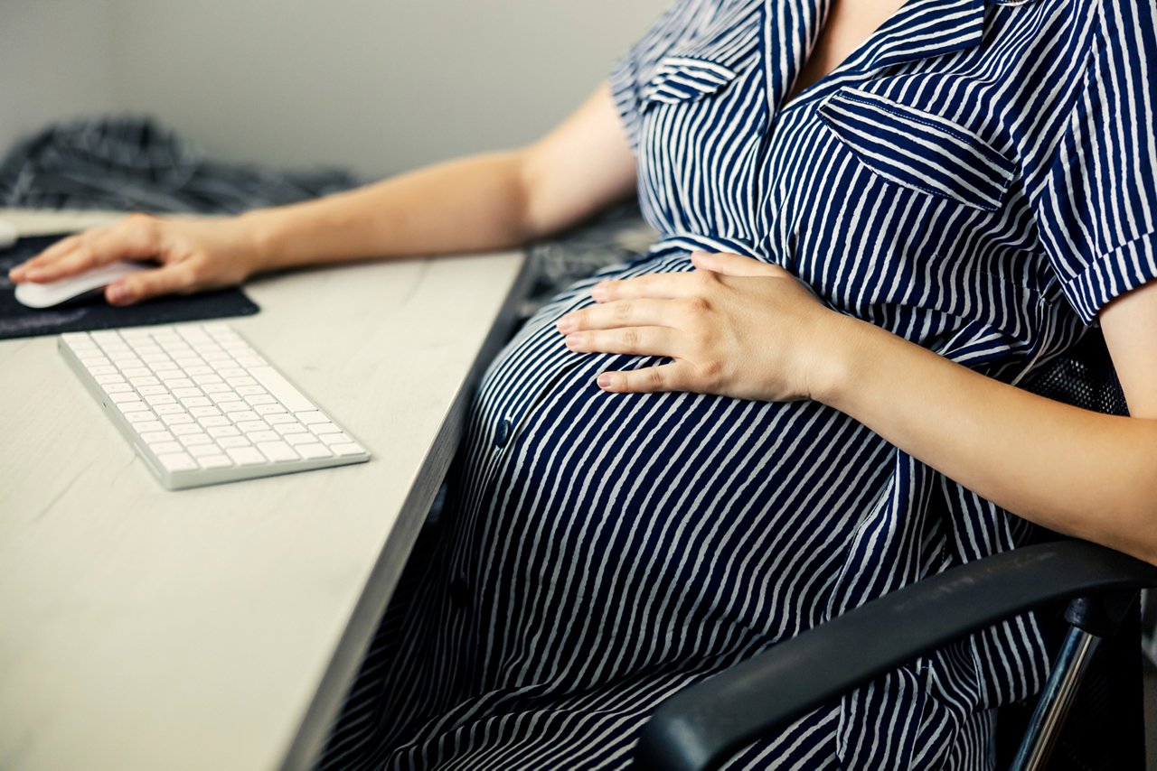 Home office and pregnant business woman wearing stripes dress, business housewife. Woman with a huge stomach sits in front of a computer with headphones in her ears and uses a mouse for online lessons