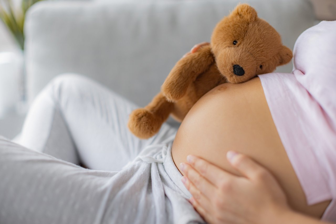 Pregnant woman caressing belly hugging teddy bear baby toy on pregnancy bump waiting for the birth of her child.