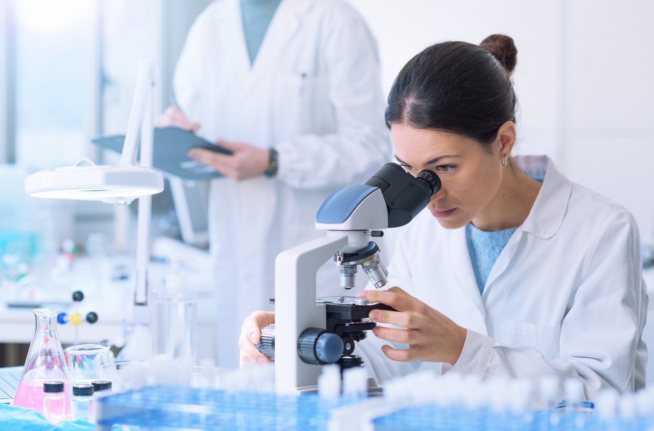 Junge Forscherin mit weißem Kittel in einem hellen Labor schaut durch ein Mikroskop