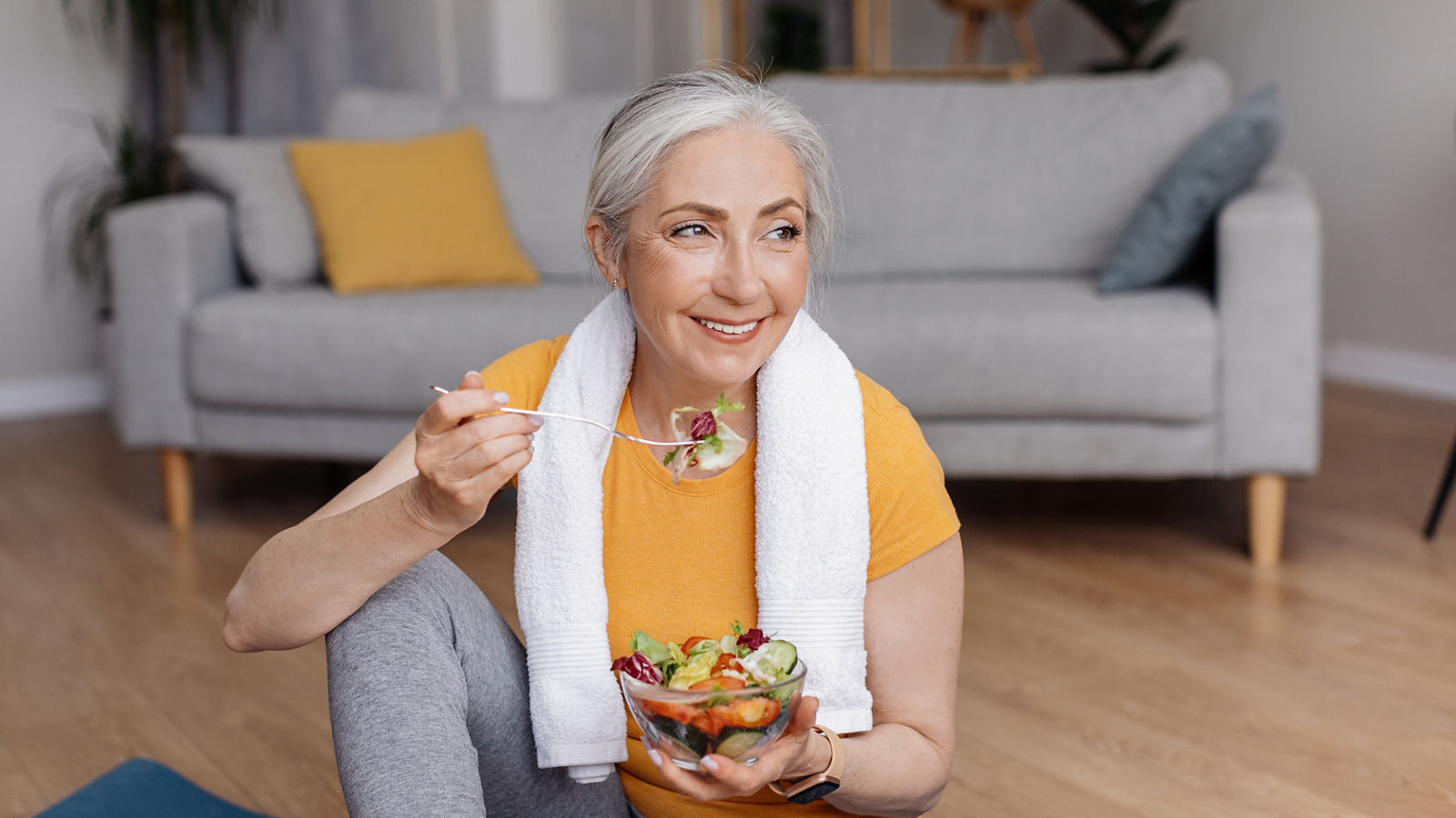 Healthy lifestyle concept. Happy senior woman eating fresh vegetable salad, sitting on yoga mat after home workout