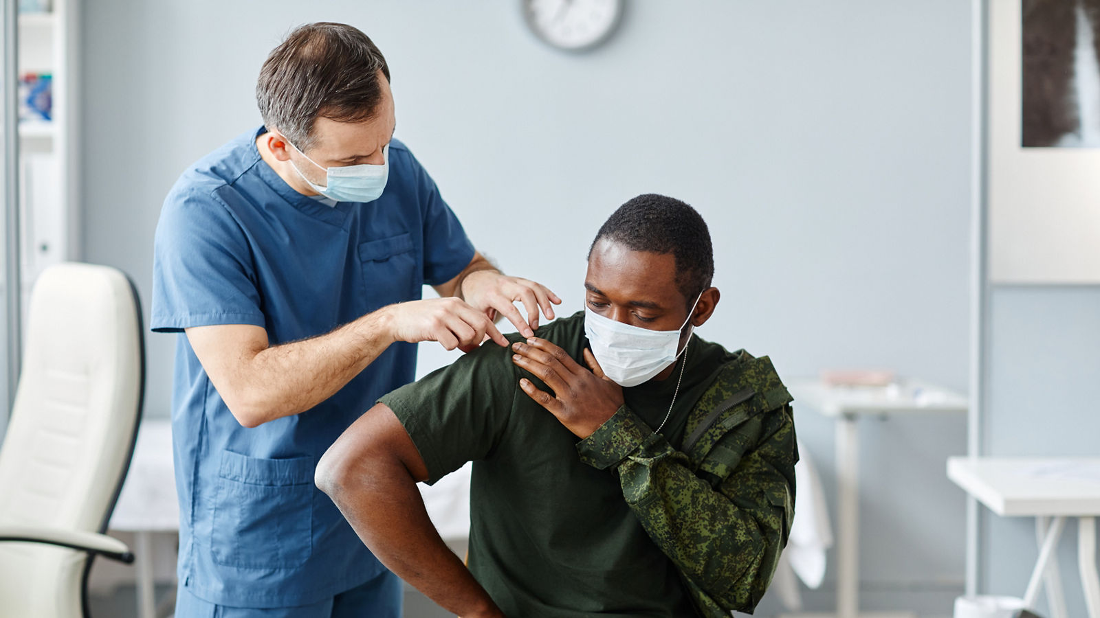 Mature doctor wearing mask and gloves palpating trapezius muscle and shoulder joint of young African American shoulder