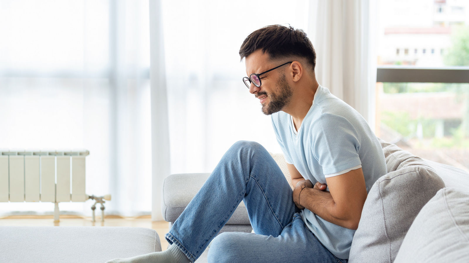 A man suffers from abdominal pain while sitting at home on the couch. Young man suffering from abdominal pain sitting on the couch at home.,A man suffers from abdominal pain while sitting at home on the c