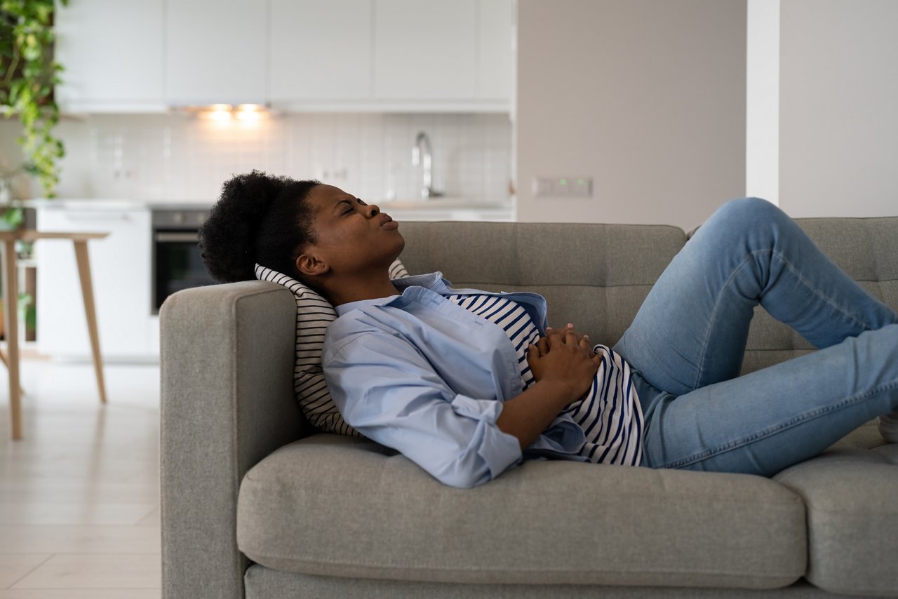 Diseased African American woman holding stomach feeling pain and anguish after alimentary poisoning or junk food. Suffering girl lies on couch needs trip to doctor gastroenterologist or nutritionist
