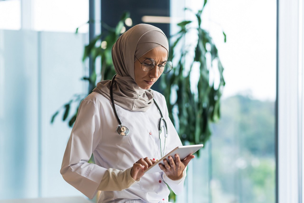 A worried and serious female doctor, an Arab, a Muslim, is standing in the corridor of the hospital, holding a tablet in her hands. Concentratedly reads, looks, there is a problem with the patient.