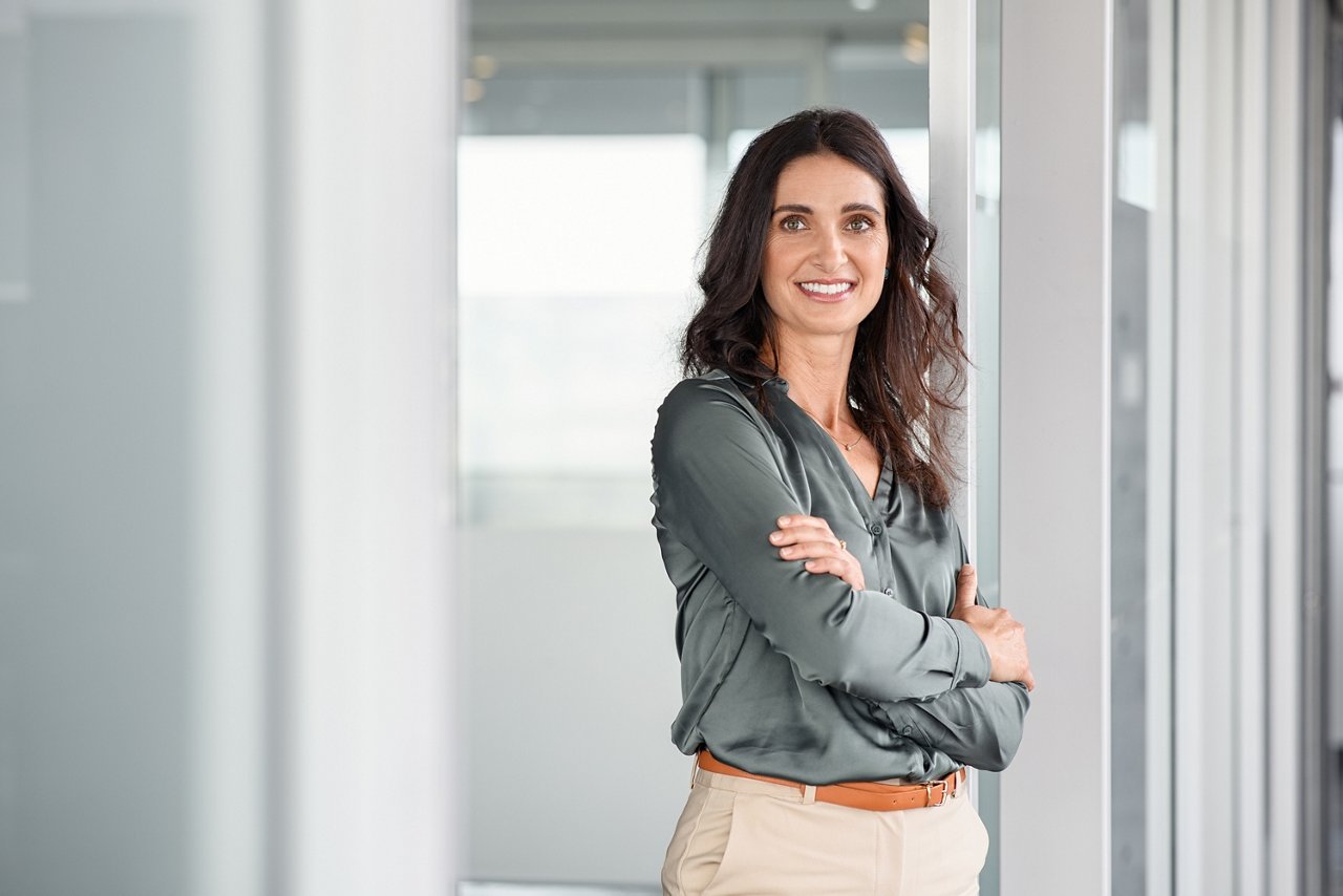 Portrait of mature business woman with arms crossed looking at camera. Happy smiling mid businesswoman standing in modern office with copy space. Confident latin woman in formalwear standing in workplace.