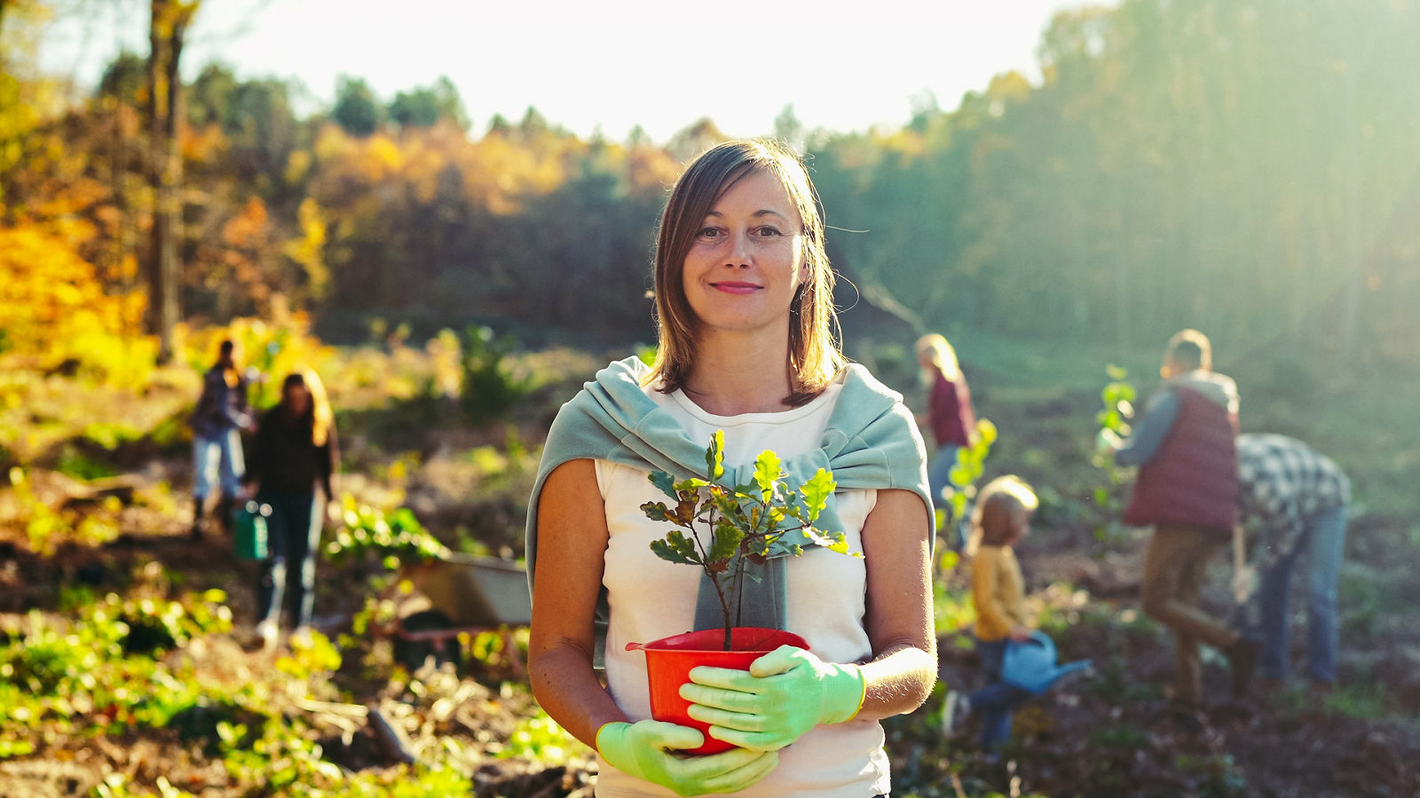 Portrait shot of young beautiful Caucasian woman standing outside with tree seedling in pot and smiling cheerfully. Pretty female eco activist working against deforestation. Green planet concept.