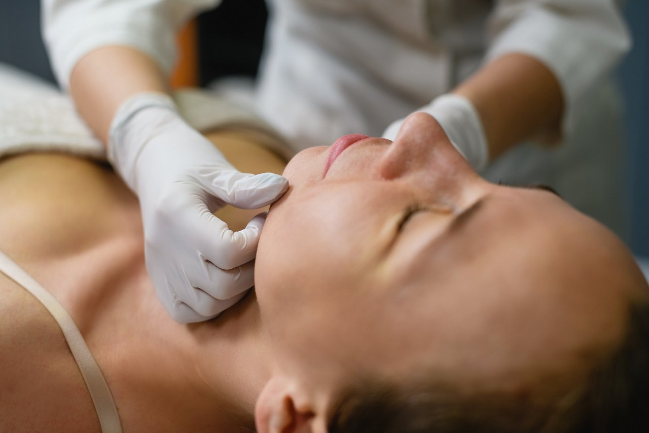Woman lay down in cosmetologist cabinet and prepare for electrolysis procedure, beautician put cream in latex gloves on skin