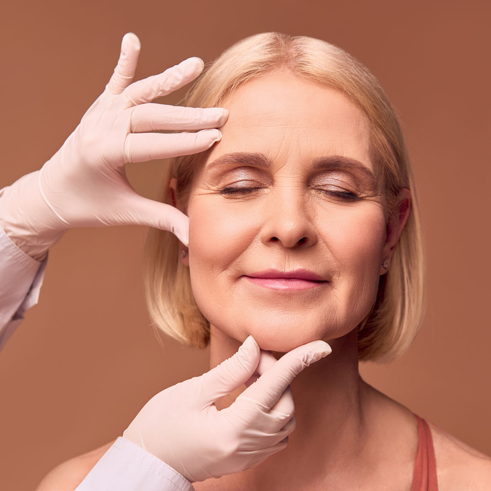 Portrait of an older adult woman with closed eyes on a beige background. Hands in white gloves and a medical gown show a face that has wrinkles.Face lift.