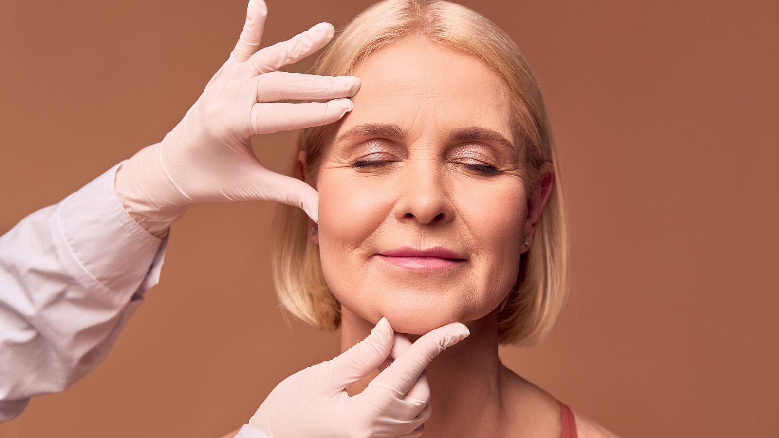 Portrait of an older adult woman with closed eyes on a beige background. Hands in white gloves and a medical gown show a face that has wrinkles.Face lift.