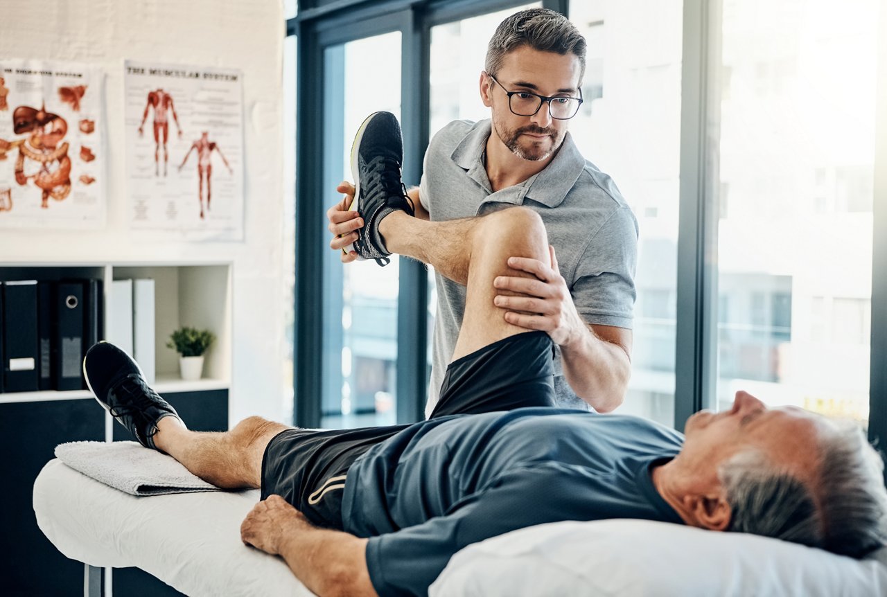 Physiotheraput bei einer bewegt Bein eines liegenden älteren Patienten bei einer Rehabilitierungsmaßnahme auf der Therapieliege