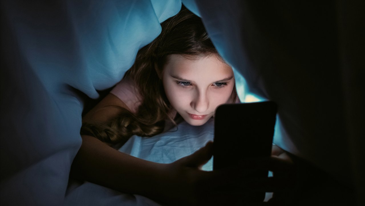 Screen time. Child insomnia. Late night chat. Internet addiction. Sleepless girl texting communicating on smartphone hiding under blanket in bed at home.