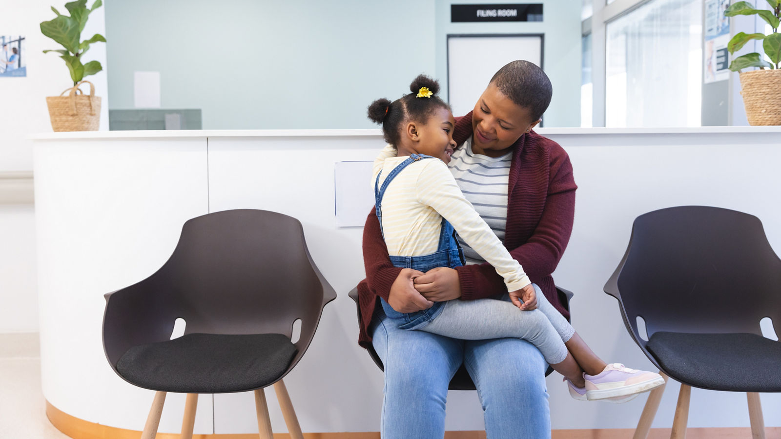 Happy african american mother and daughter sitting in waiting room at hospital, copy space