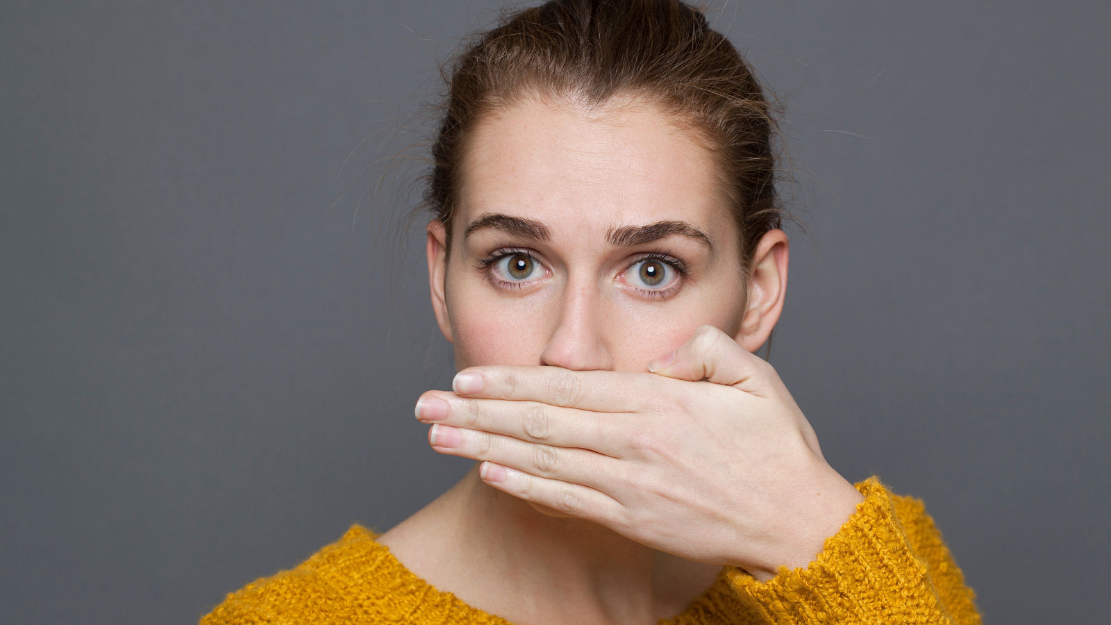 negative feelings concept - portrait of surprised beautiful 20s girl covering her mouth for bad breath or taboo,studio shot on gray background