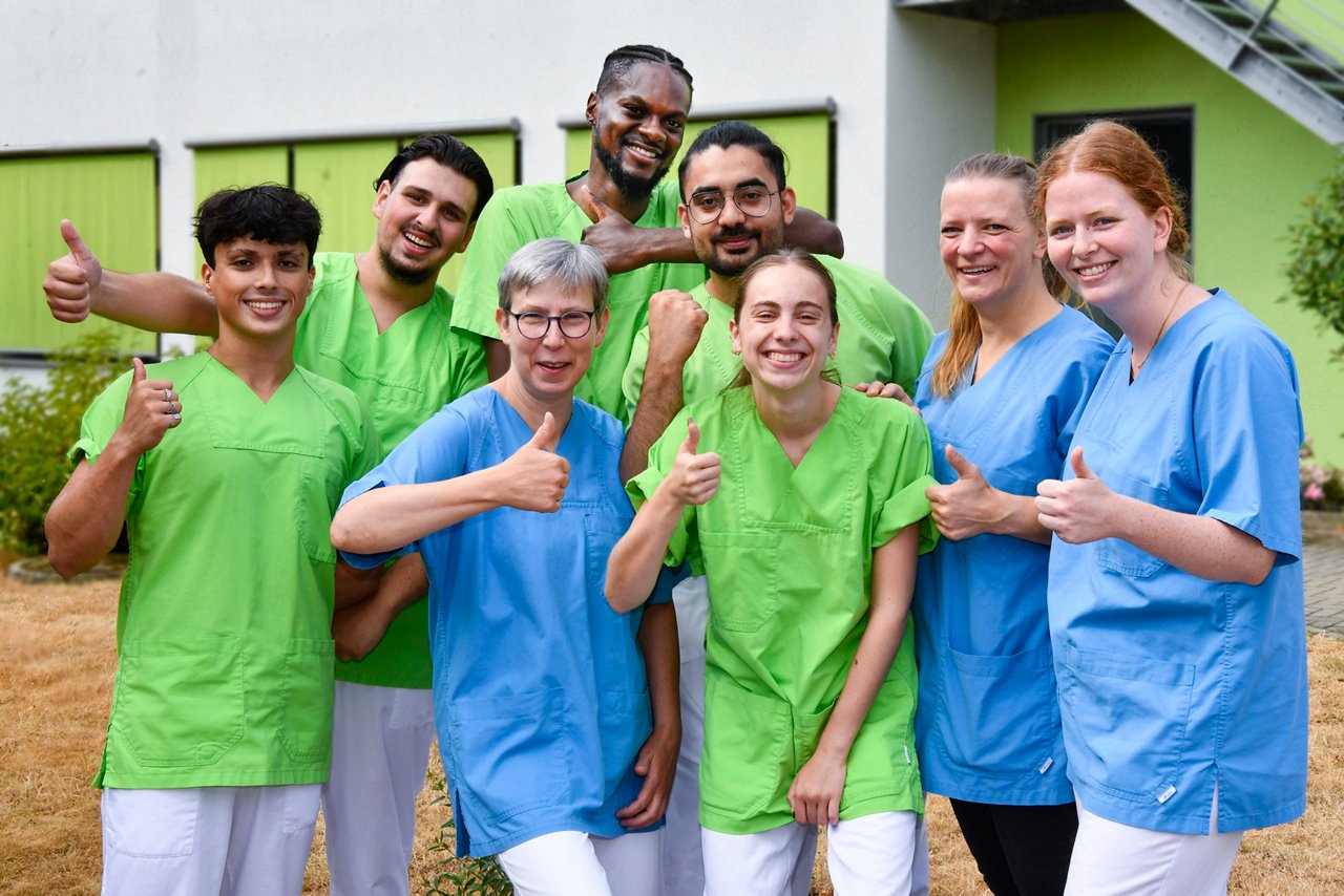 Azubi-Gruppe lacht, hat Spaß und zeigt Daumen hoch für die Pflege-Ausbildung. 