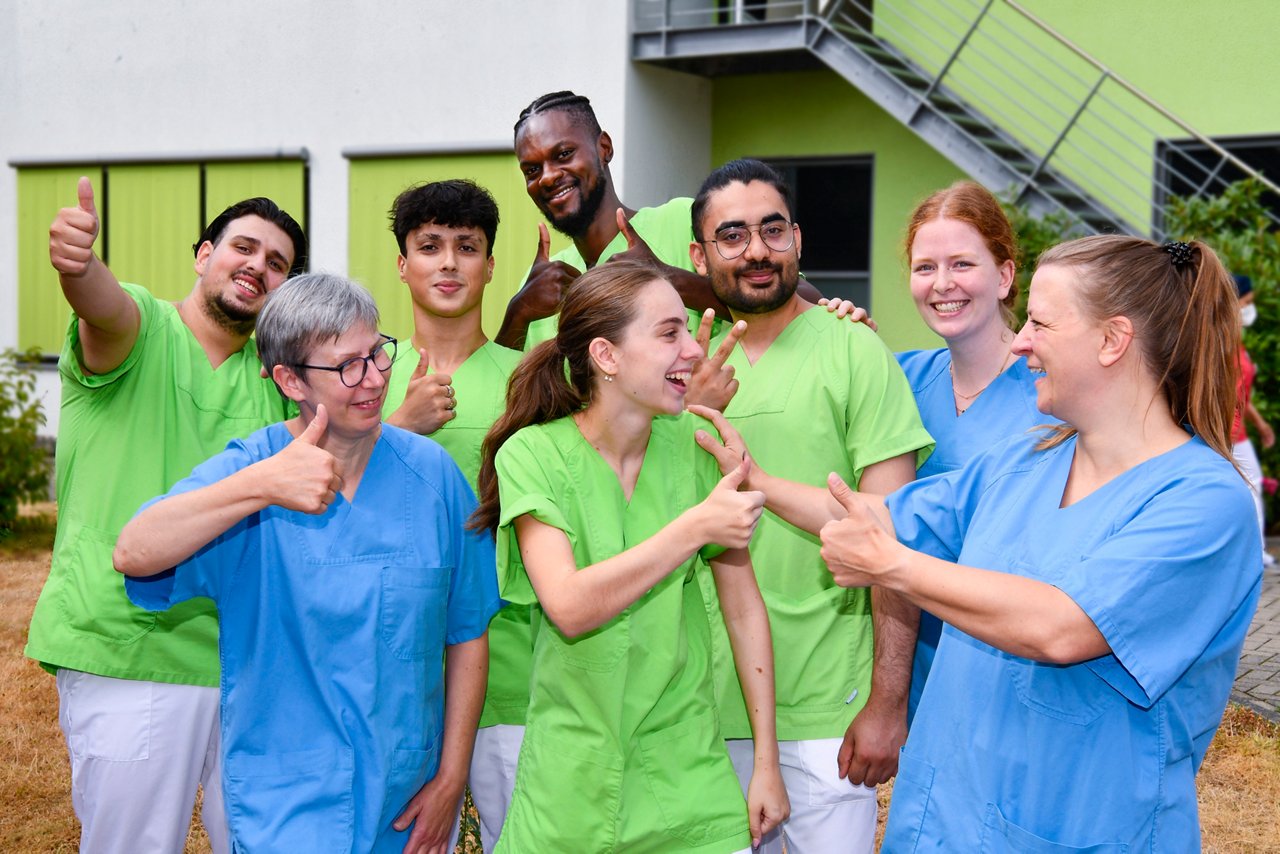 Azubi-Gruppe lacht, hat Spaß und zeigt Daumen hoch für die Pflege-Ausbildung. 